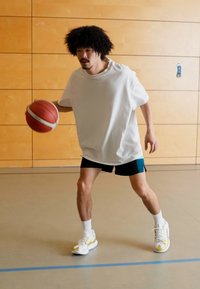 Un homme athlétique portant un t-shirt blanc ample et un short noir, dribblant un ballon de basketball dans une salle de sport avec des murs en bois.