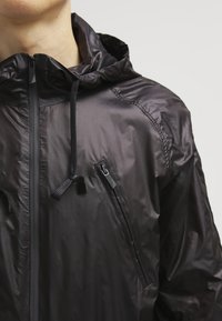 Veste noire légère à capuche avec une poche zippée, des cordons élastiques et une texture lisse. Présente un design épuré et minimaliste.