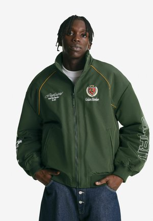 Green bomber jacket with ribbed cuffs, yellow accents, logo patches, and a front zipper. Worn with blue denim jeans.