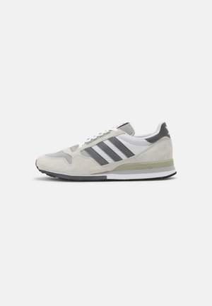 Beige en grijze Adidas sneaker met witte veters, drie donkergrijze strepen en logo op de hiel en zool, zijaanzicht op witte achtergrond.
