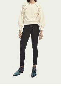 Felpa crema con maniche a sbuffo e cuciture decorative, abbinata a jeans skinny neri e stivaletti teal con accenti metallici.