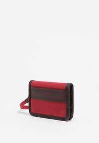 Portafoglio rosso in tessuto con un accento in pelle marrone. Presenta una chiusura con zip, una tracolla in tessuto e una forma rettangolare con cuciture a contrasto.
