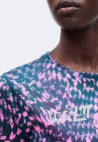 Κοντινό πλάνο ενός ατόμου που φοράει ένα ροζ και σκούρο πράσινο πουκάμισο με το κείμενο «tcs L17» στο στήθος.