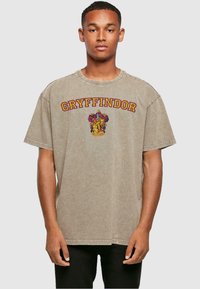 Camiseta de gran tamaño beige hecha de algodón. Presenta el texto "GRYFFINDOR" en color marrón y un diseño de escudo colorido en la parte delantera. Mangas cortas.