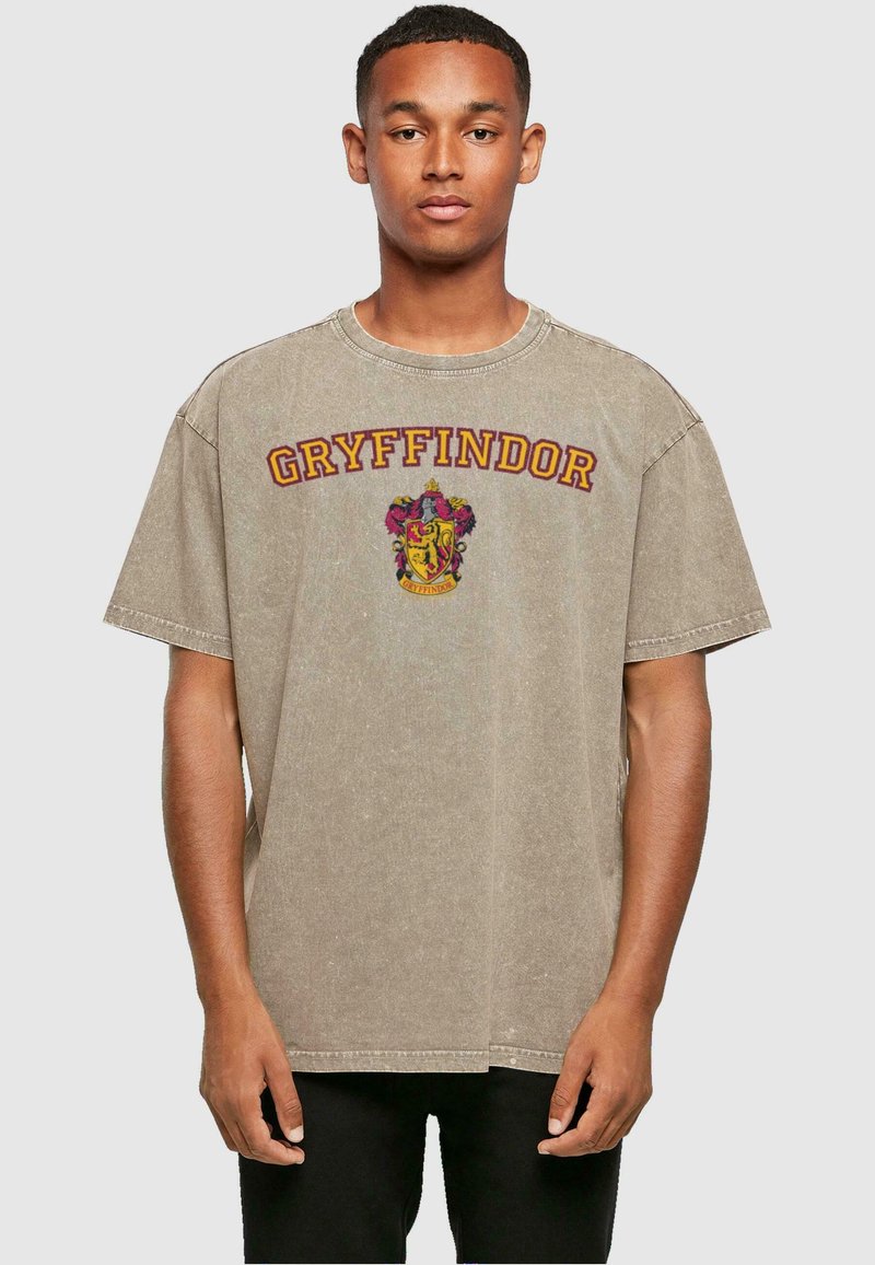 Camiseta de gran tamaño beige hecha de algodón. Presenta el texto "GRYFFINDOR" en color marrón y un diseño de escudo colorido en la parte delantera. Mangas cortas.