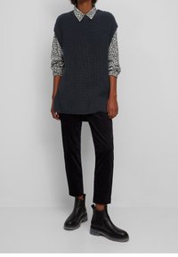 Tunique gris foncé en tricot avec une texture côtelée, superposée à une chemise blanche ornée d'un motif floral noir, associée à un pantalon en velours côtelé noir et des bottines.