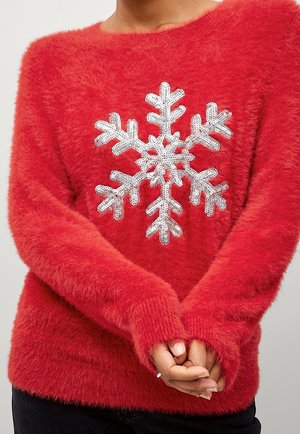 Czerwony, puszysty sweter z dużym białym, cekinowym wzorem śnieżynki, noszony przez osobę ze splecionymi rękami z przodu.