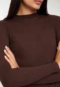 Kobieta z założonymi rękami, ubrana w brązowy, prążkowany, długi rękaw o wysokim dekolcie, z widoczną częścią twarzy i rąk.