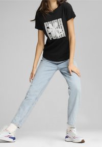 Schwarzes T-Shirt mit einem grafischen Logo-Design. Kombiniert mit hellblauen Jeans und weißen Sneakers mit lila und grauen Akzenten.