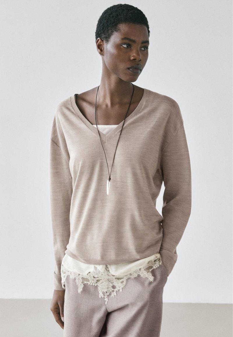 Femme aux cheveux courts et bouclés portant un pull beige à col en V, un top blanc bordé de dentelle, un pantalon mauve clair et un long collier avec pendentif.