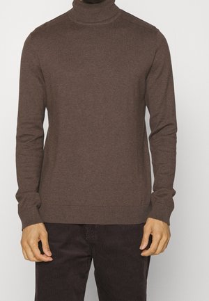 Homme portant un pull col roulé marron ajusté et un pantalon en velours côtelé marron foncé, debout devant un fond uni.