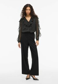 Blusa nera a pois con colletto arricciato e maniche lunghe trasparenti, abbinata a pantaloni neri a vita alta e a gamba larga e tacchi a punta.