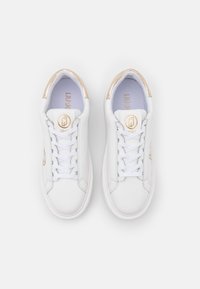 Valkoiset nahkakengät, joissa on sileä pinta, kultaiset yksityiskohdat kantapäässä ja valkoinen nauhakiinnitys edessä. Logo näkyy sivulla ja kielessä.