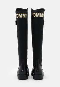 Mustat polven korkeudelle ulottuvat saappaat, joissa on kangas yläosa, jossa lukee "TOMMY" kultanaisella ompeleella, sekä sileä nahka alaosa ja teksturoitu pohja.
