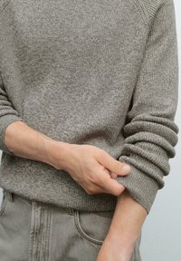 Bruin gebreid sweater met opgerolde manchetten, die een gestructureerd patroon en ribbelafwerking bij de hals en zoom laten zien. G gedragen met grijze broek.