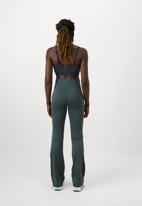 Temno siva športna modrček z teksturirano tkanino in nastavljivimi naramnicami, v kombinaciji s prilegajočimi, širokimi zelenimi trening hlačami iz gladkega materiala.