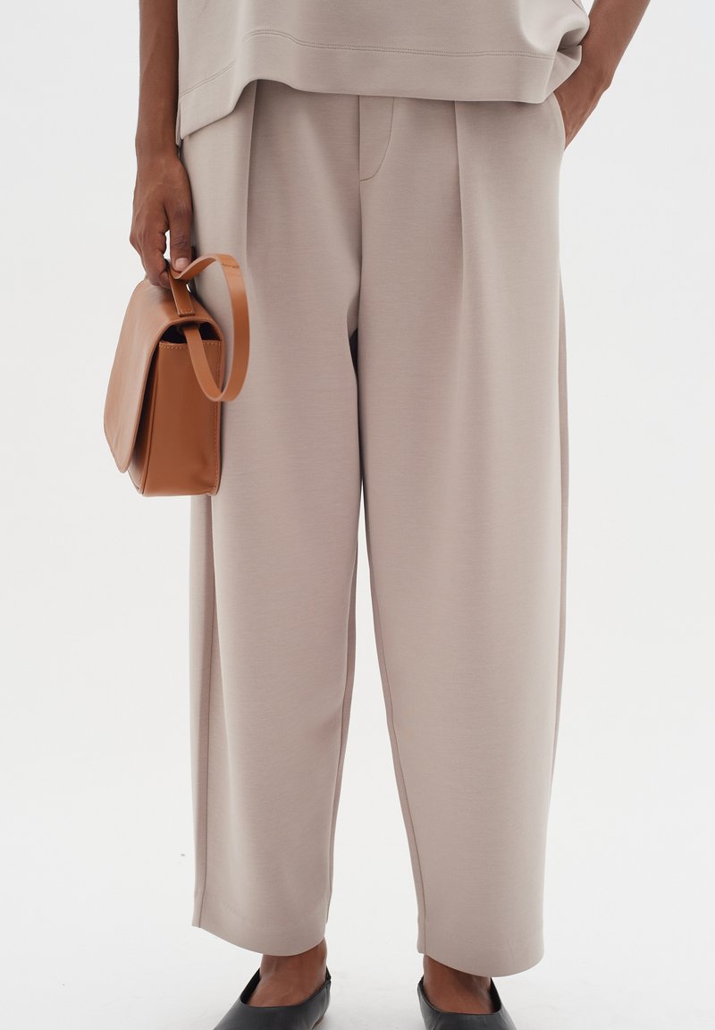 Beige wide-leg trousers with front pleats, smooth texture, paired with a brown handbag and black flat shoes.