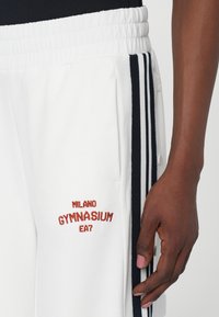 Baltas sporta bikses ar tumši zilām svītrām sānos, ar izšūtu "Milano Gymnasium EA7" logotipu sarkanā krāsā. Elastīga josta un sānu kabatas.