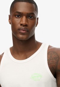 Camiseta sin mangas blanca con un logo verde de "SuperDry". La tela parece suave y la camiseta presenta un cuello redondo.