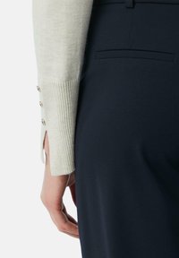 Beige gebreide trui met een ribbelstructuur en knoopdetails, gecombineerd met marineblauwe stoffen broek met een achterzakdetail.