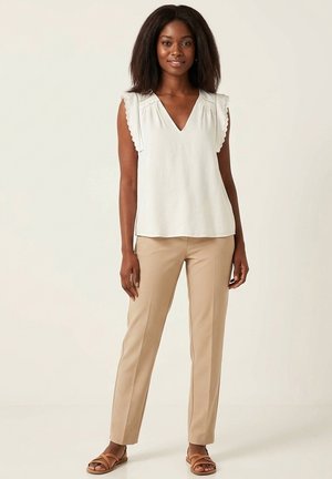 Femme debout portant un chemisier blanc sans manches avec des bords festonnés, un pantalon beige et des sandales plates marron sur un fond uni.