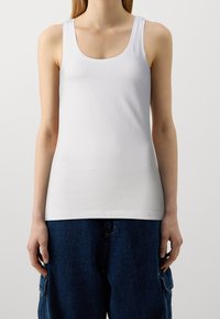 Mujer vistiendo una camiseta blanca ajustada y pantalones cargo oscuros sueltos, de pie contra un fondo claro y sencillo.