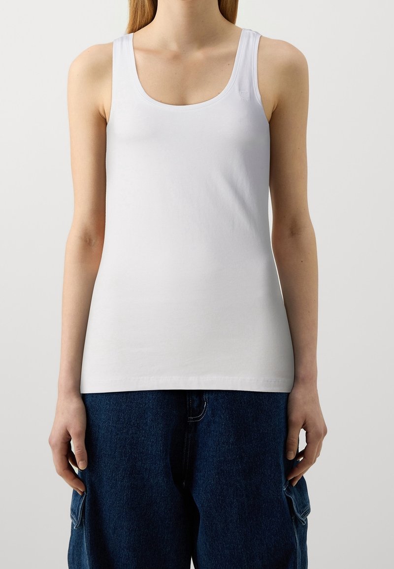 Mujer vistiendo una camiseta blanca ajustada y pantalones cargo oscuros sueltos, de pie contra un fondo claro y sencillo.