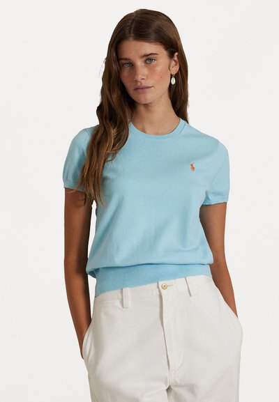 Maglione a maniche corte di cotone azzurro chiaro con scollo rotondo e orlo a coste, caratterizzato da un logo arancione sul petto. Abbinato a pantaloni bianchi.