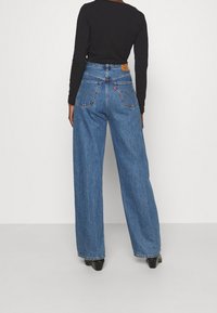 Bredbeniga blå denimjeans med hög midja. Har två bakfickor med sömnadsdetaljer. Svart långärmad topp delvis synlig.