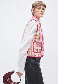 Ένα πολύχρωμο γιλέκο με σχέδια σε ροζ, κόκκινο και κίτρινο, με κουμπιά και τσέπη. Φορεμένο πάνω από λευκό πουκάμισο, συνδυασμένο με σκούρο τζιν παντελόνι και μπορντό τσάντα.