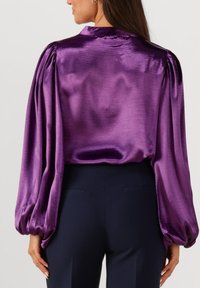 Paarse satijnen blouse met een zachte textuur, oversized puffmouwen en een hoge halslijn, gedragen over donkere op maat gemaakte broeken.