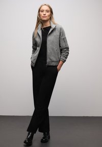 Šedá bomber bunda s texturou, stojáčkovým límcem, předním zipem a bočními kapsami, doplněná o černý top a černé zúžené kalhoty.