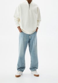 Felpa bianca con zip, vestibilità rilassata, abbinata a jeans a gamba larga blu chiaro e sneakers bianche con dettagli marroni. Tessuto liscio in tutto.
