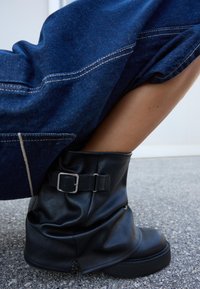 Zwarte leren enkelboot met een gevouwen ontwerp, zilveren gespdetail en een dikke zool. Denimhuid en huid gedeeltelijk zichtbaar op de achtergrond.