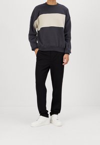 Sweter w ciemnoszarym kolorze z kremowym poziomym paskiem, zestawiony z czarnymi spodniami i białymi sneakerami. Tkanina wydaje się miękka i casualowa.