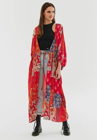 Červený kimono s vzorem, ozdobený modrými a oranžovými akcenty, s dlouhými rukávy, otevřeným předním dílem a volným střihem, v kombinaci s černým topem a šedými džíny.