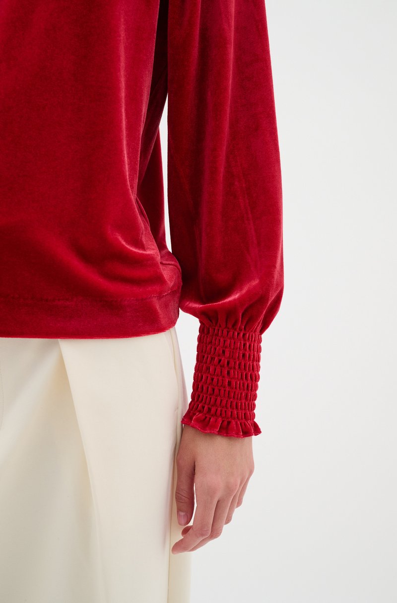 Rotes Samtoberteil mit langen Ärmeln und gerafften, gesteppten Manschetten. Weiche Textur, glatte Oberfläche, figurbetonte Silhouette und kontrastierende cremefarbene Hosen.