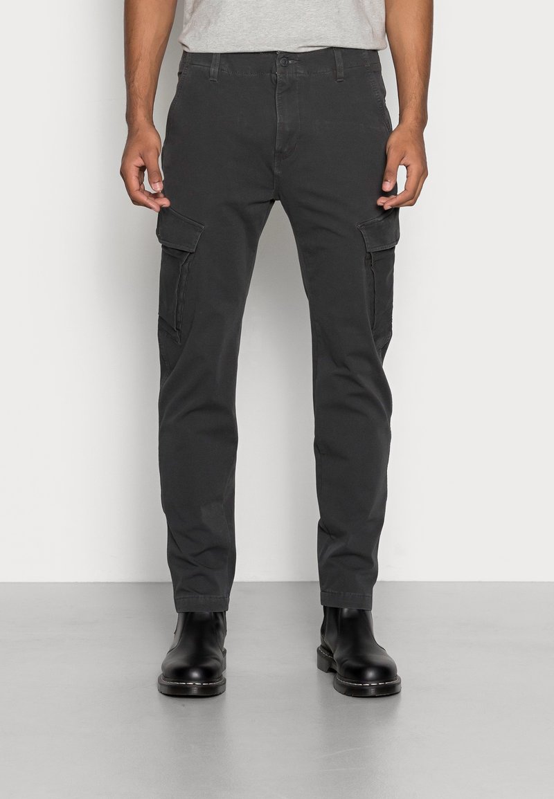 Homme portant un pantalon cargo noir avec des poches à rabat latérales et des bottes en cuir noir, debout devant un fond clair uni.