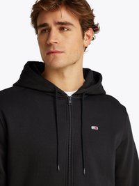 Sweat à capuche noir zippé en tissu doux, avec une fermeture éclair sur le devant, des cordons de serrage ajustables et un petit logo sur la poitrine gauche.