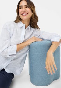 Striped light blue shirt with rolled sleeves, paired with a cylindrical, textured light blue ottoman. Silver bracelet on wrist.
