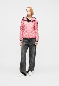 Γυναίκα που στέκεται φορώντας ροζ καπιτονέ τζάκετ, μαύρο πουκάμισο, άνετα σκουρόγκρι τζιν και λευκά αθλητικά παπούτσια μπροστά σε απλό φόντο.