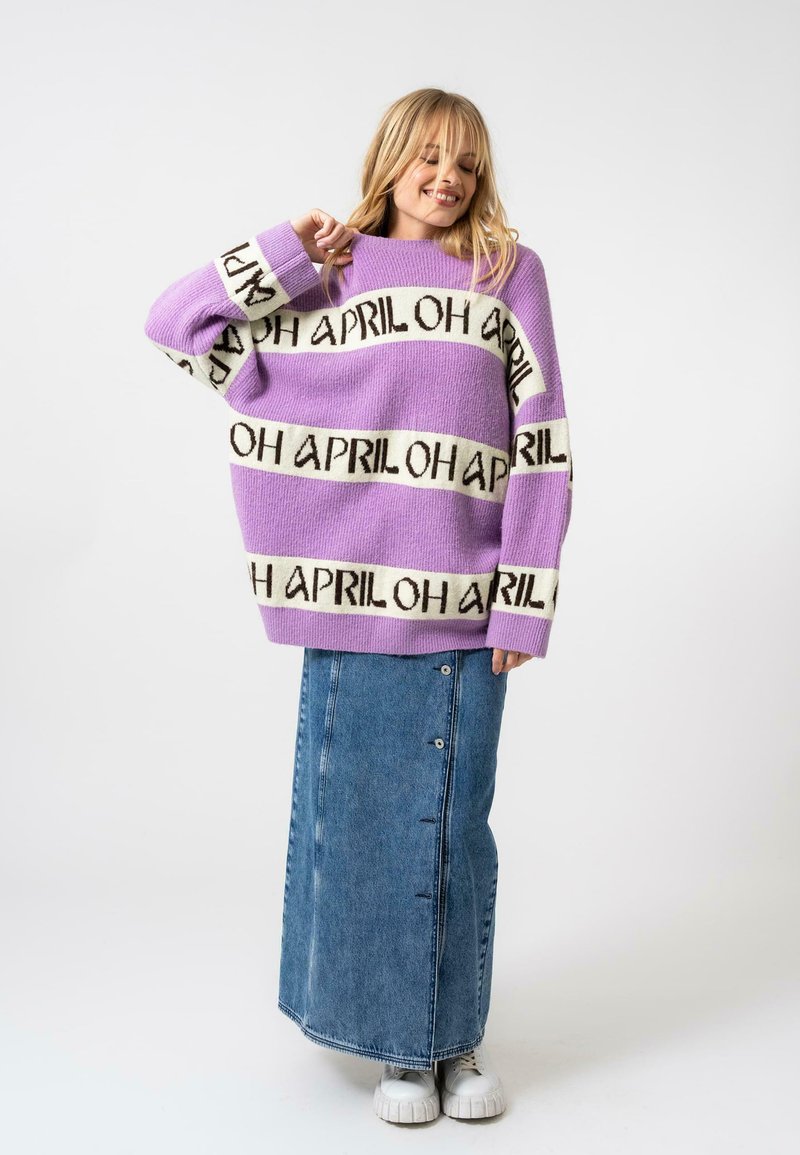 Un pull en maille violet avec des bandes de texte "APRIL" en noir et blanc ; associé à une jupe maxi en denim avec des boutons sur le devant et des baskets blanches à plateforme.