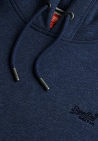 Sudadera con capucha azul marino de tejido texturizado, que presenta un logotipo y cordones con puntas de marca. Detalle sutil de costura visible.