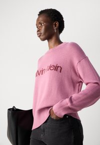 Donna che indossa un maglione rosa di Calvin Klein, jeans scuri, con una grande borsa nera, che guarda di lato su uno sfondo semplice.