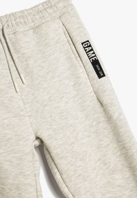 Lys grå joggerbukser lavet af blødt, struktureret stof med en elastisk talje. Har et sort mærke med ordet "GAME" og en snor.