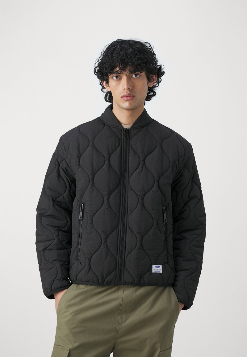 Czarna pikowana kurtka z wysokim kołnierzem, zapinaną na zamek z przodu i dwoma bocznymi kieszeniami. Posiada falisty wzór oraz małą metkę marki u dołu.