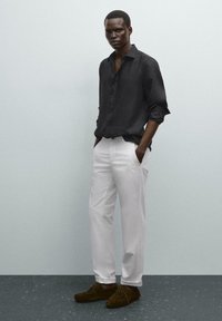 Schwarzes Leinenhemd, weiße Hose und braune Wildlederschuhe. Das Hemd hat einen Knopfverschluss; die Hose ist locker sitzend und verfügt über Taschen.