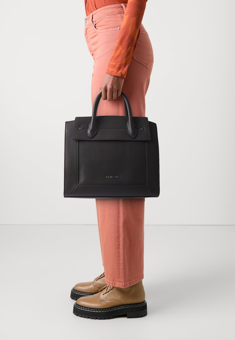 Borsa a tracolla in pelle nera con design strutturato, manici piatti, minimal hardware e logo sottile. Abbinata a pantaloni rosa pastello e scarpe marroni.
