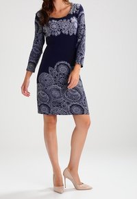 Marineblaues Kleid mit Dreiviertelärmeln, verziert mit weißen filigranen Blumenmustern und einem Rundhalsausschnitt, kombiniert mit beigen High Heels.
