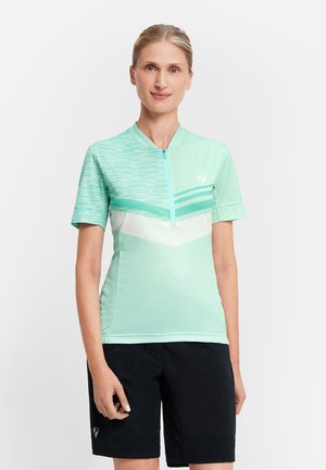 Frau trägt ein mintgrünes Radtrikot mit kurzen Ärmeln und Reißverschluss sowie schwarze Shorts, steht vor einem schlichten weißen Hintergrund.